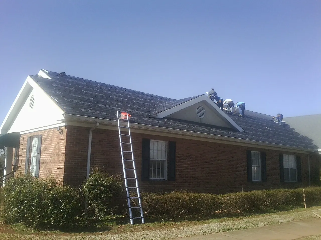 Crew tearing shingles off a brick ranch roof with a ladder set — Emergency Roof Repair work by our crew in Nelliston