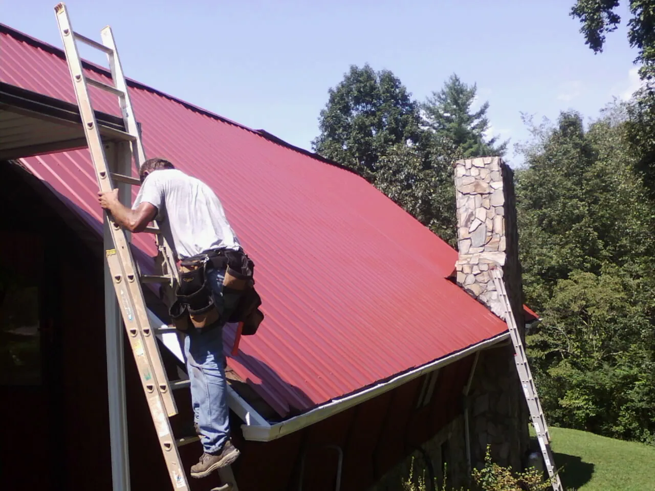 Roofer on a ladder beside a red metal roof and stone chimney — Soffit & Fascia Repair by Roofing Brothers in Nelliston